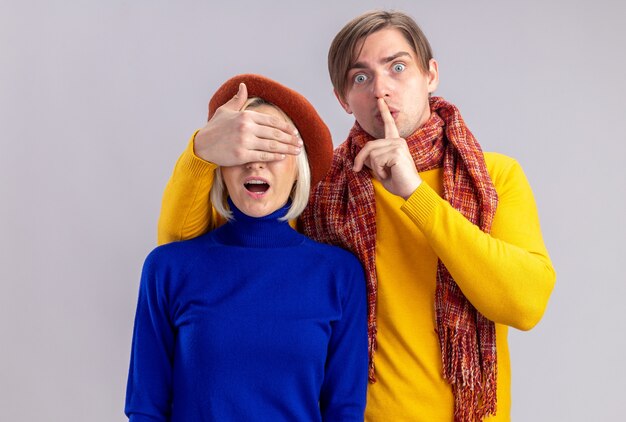 Anxious handsome slavic man with scarf around his neck doing silence gesture and closing eyes of pretty blonde woman with beret on valentine's day 