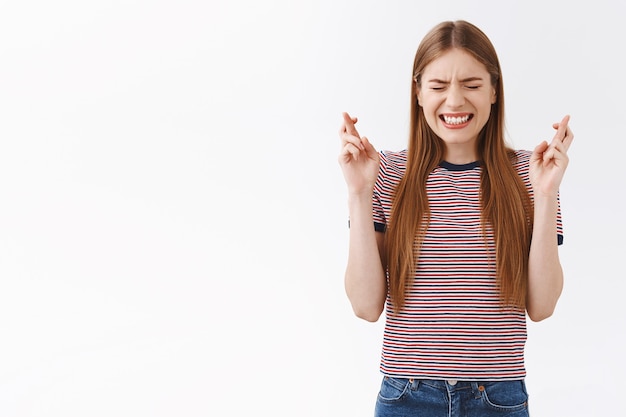 Anxious hopeful young fair-haired caucasian girl in striped t-shirt praying, clench teeth nervously close eyes and cross fingers good luck, anticipating something important, nervously awaiting