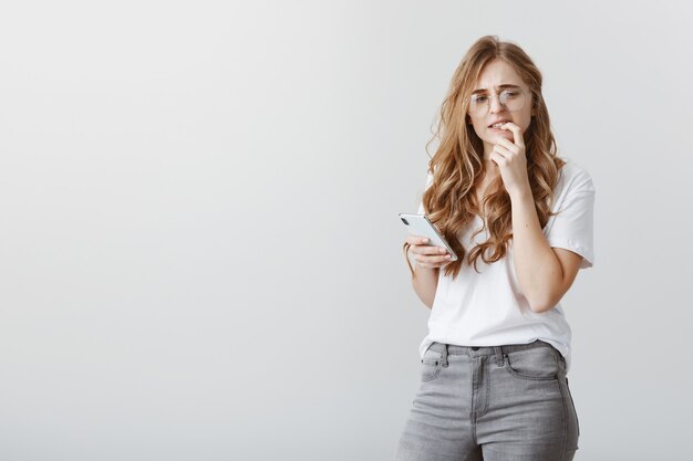 Anxious and insecure in herself. Worried good-looking european woman with blond hair in trendy glasses, standing half-turned holding smartphone, biting finger while thinking what to answer, hesitating