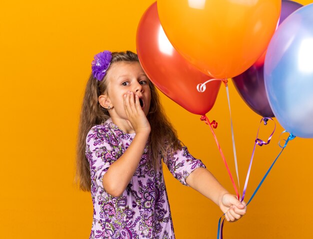 ヘリウム気球を保持し、コピースペースでオレンジ色の壁に隔離された顔に手を置く気になる小さなブロンドの女の子
