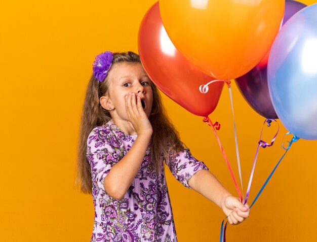 anxious little blonde girl holding helium balloons and putting hand on face isolated on orange wall with copy space