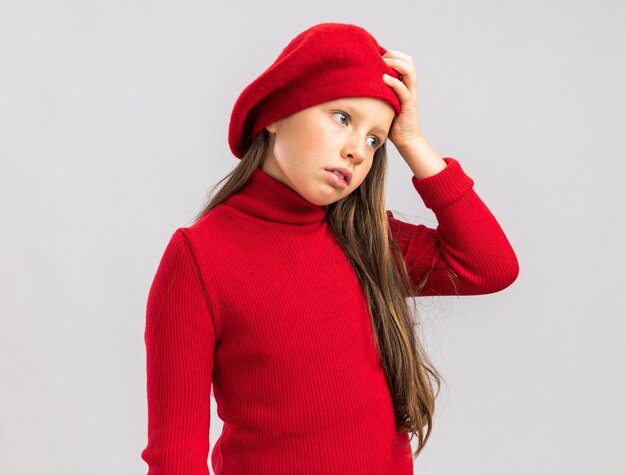 Anxious little blonde girl standing in profile view wearing red beret keeping hand on head looking side isolated on white wall with copy space