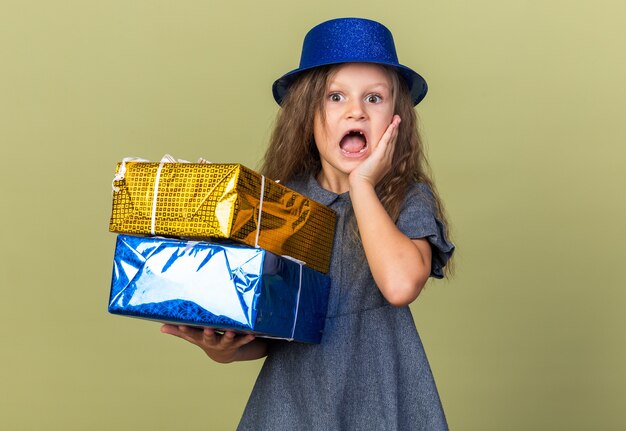 anxious little blonde girl with blue party hat putting hand on face and holding gift boxes isolated on olive green wall with copy space