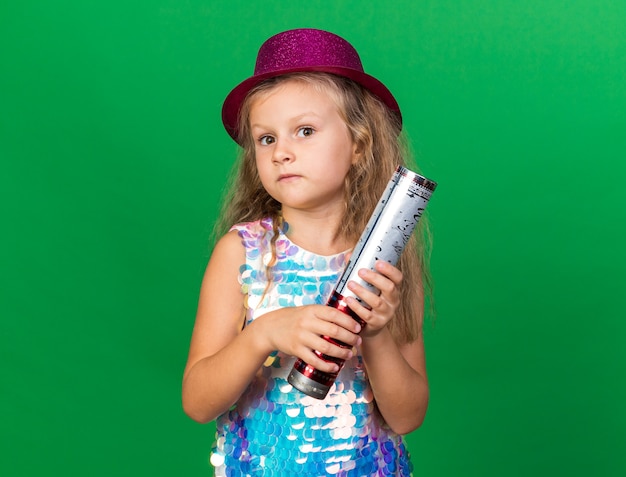 anxious little blonde girl with purple party hat holding confetti cannon isolated on green wall with copy space