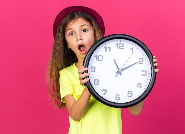 anxious little caucasian girl with purple party hat holding clock isolated on pink wall with copy space