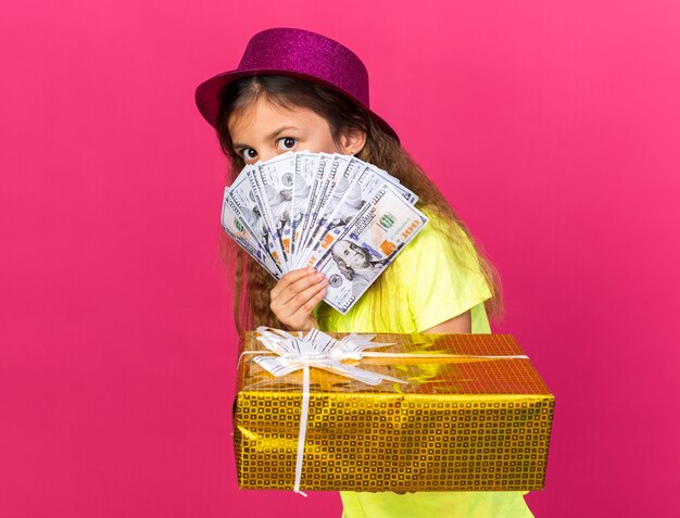 anxious little caucasian girl with purple party hat holding gift box and money isolated on pink wall with copy space