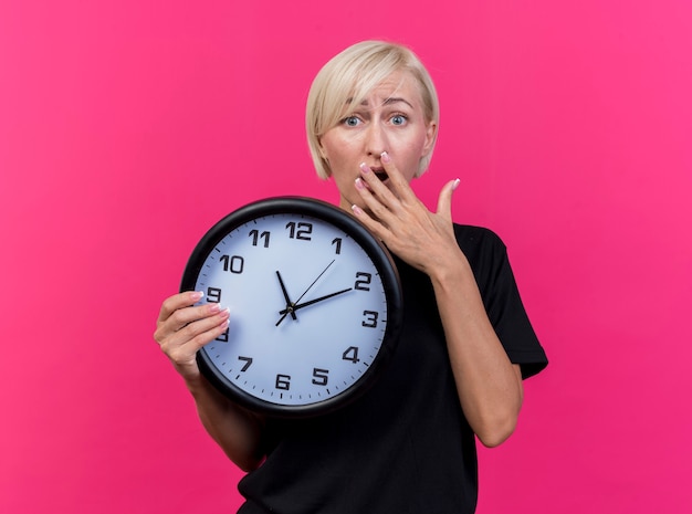 Free photo anxious middle-aged blonde slavic woman holding clock  keeping hand on mouth isolated on crimson wall