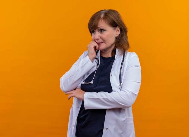 의료 가운 및 복사 공간이 격리 된 주황색 벽에 턱에 손을 넣어 청진기를 착용하는 불안 중년 여성 의사