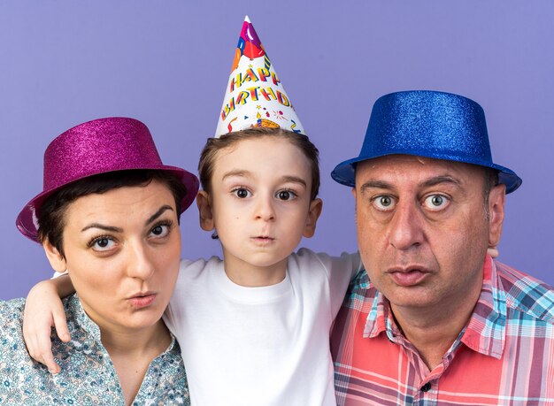 anxious mother and father with party hats standing with their son isolated on purple wall with copy space