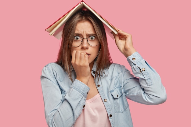 Anxious nervous Caucasian female bites finger nails, holds book above head, worries before passing exam, poses against pink backgroud. Student looks nervously. People and education concept