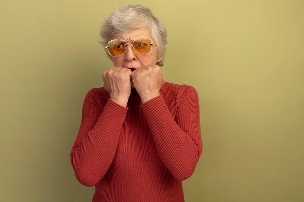 Free photo anxious old woman wearing red turtleneck sweater and sunglasses looking at side keeping hands on mouth isolated on olive green wall with copy space