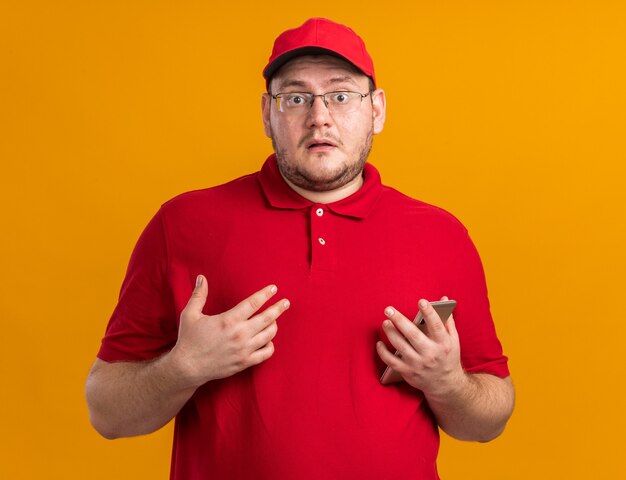 anxious overweight young deliveryman in optical glasses holding phone isolated on orange wall with copy space