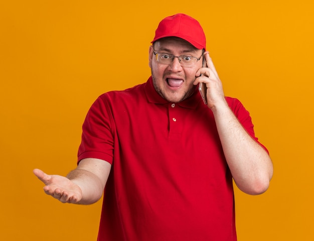 Anxious overweight young deliveryman in optical glasses talking on phone isolated on orange wall with copy space