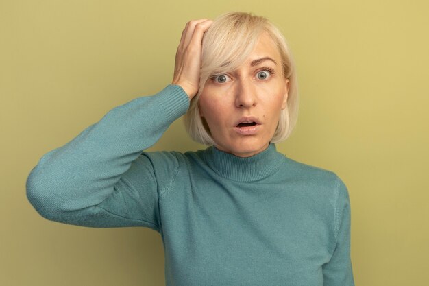 Anxious pretty blonde slavic woman puts hand on head and looks at camera on olive green