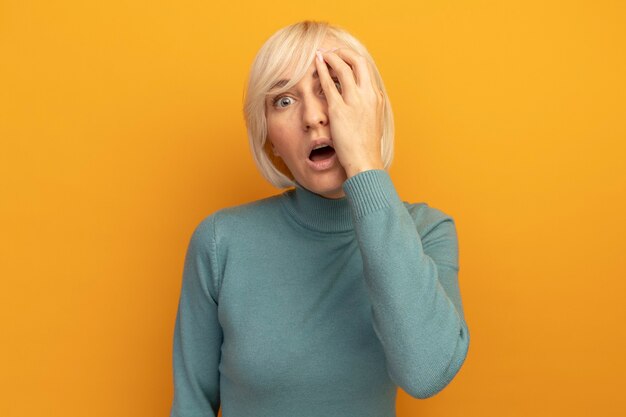 Anxious pretty blonde slavic woman putting hand on face and looking at front isolated on orange wall