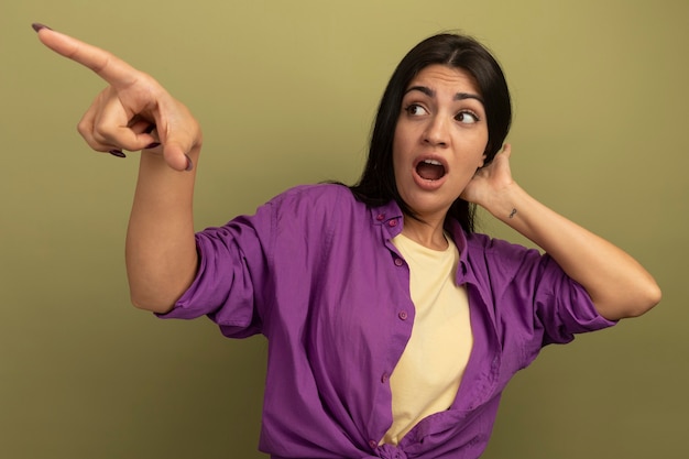 La ragazza caucasica piuttosto mora ansiosa mette la mano sulla testa dietro guardando e indicando a lato su verde oliva