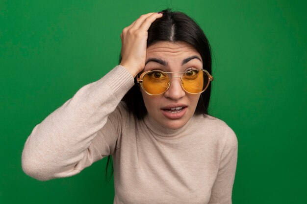 Anxious pretty brunette caucasian girl in sun glasses puts hand on head on green