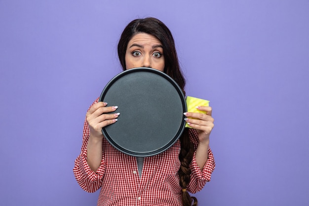 Foto gratuita donna caucasica ansiosa delle pulizie che tiene piatto e spugna e guarda