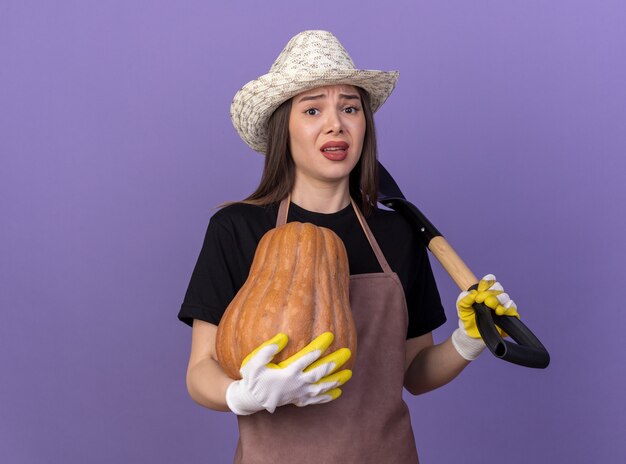 Anxious pretty caucasian female gardener wearing gardening hat and gloves holding pumpkin and spade on shoulder isolated on purple wall with copy space