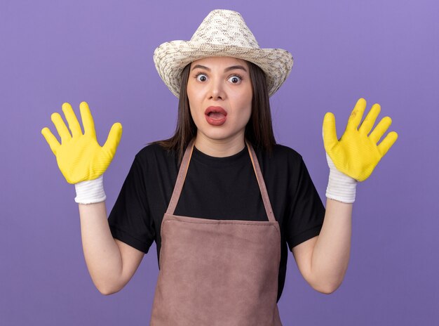 コピースペースと紫色の壁に隔離された上げられた手で立っているガーデニングの帽子と手袋を身に着けている気になるかなり白人女性の庭師