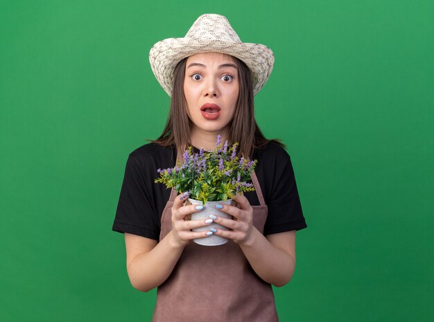 Anxious pretty caucasian female gardener wearing gardening hat holding flowerpot on green