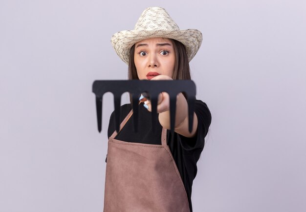 Anxious pretty caucasian female gardener wearing gardening hat holding out rake isolated on white wall with copy space