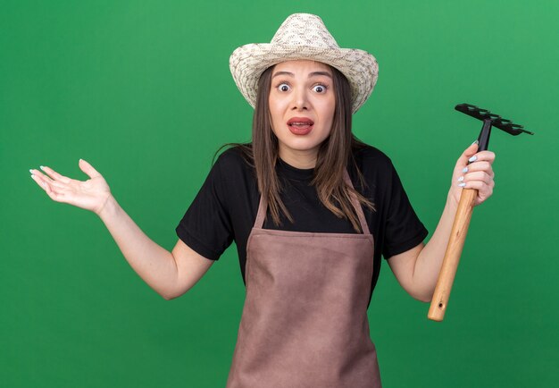 Anxious pretty caucasian female gardener wearing gardening hat holding rake and keeping hand open isolated on green wall with copy space