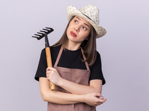 Anxious pretty caucasian female gardener wearing gardening hat holding rake and looking at side