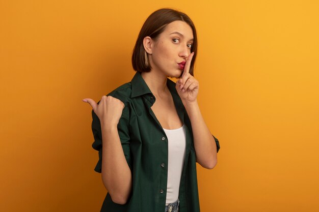 Anxious pretty caucasian woman doing silence gesture and points at side on orange