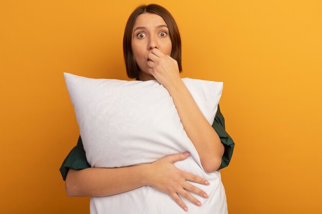 Anxious pretty caucasian woman holds pillow and bites nails on orange
