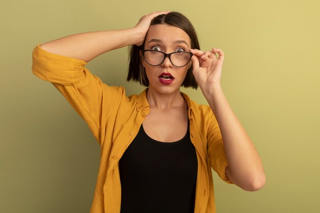 Anxious pretty caucasian woman puts hand on head and looks at camera through optical glasses on olive green