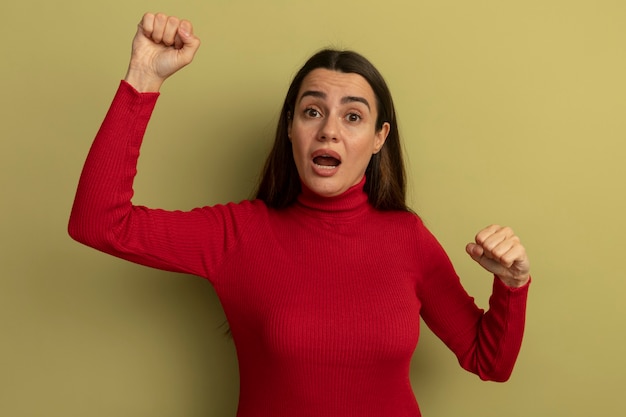 Anxious pretty caucasian woman raising fists up on olive green