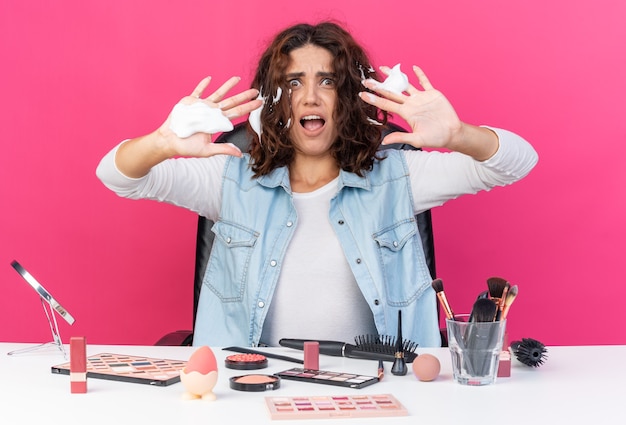 Foto gratuita donna abbastanza caucasica ansiosa seduta al tavolo con strumenti per il trucco applicando mousse per capelli e tenendo le mani aperte isolate sulla parete rosa con spazio di copia