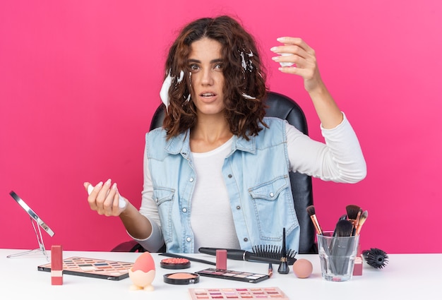Тревожная симпатичная кавказская женщина сидит за столом с инструментами для макияжа и наносит мусс для волос