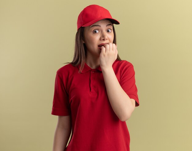 Anxious pretty delivery woman in uniform bites nails isolated on olive green wall with copy space