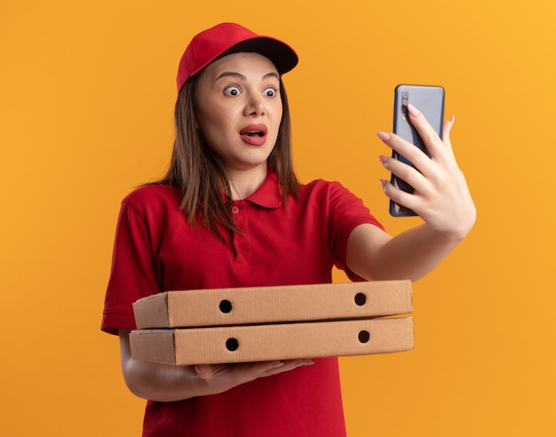Anxious pretty delivery woman in uniform holds pizza boxes and looks at phone on orange