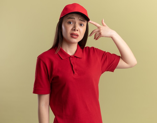 Anxious pretty delivery woman in uniform points at cap on olive green