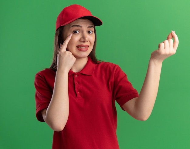 Anxious pretty delivery woman in uniform puts finger on eyelid and pretends to hold something on green