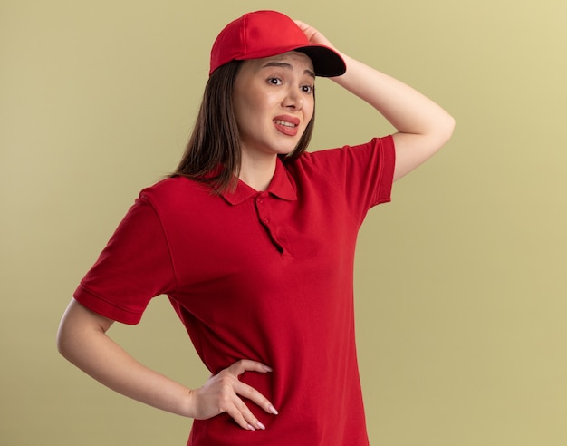Anxious pretty delivery woman in uniform puts hand on head and looks at side isolated on olive green wall with copy space