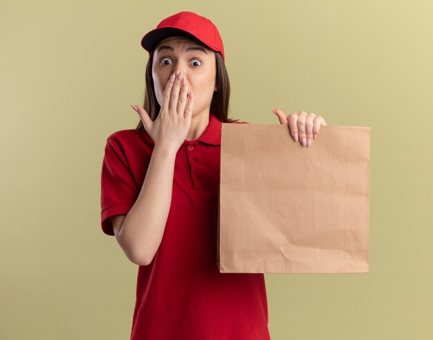 Обеспокоенная симпатичная доставщица в униформе кладет руку на рот и держит бумажный пакет на оливково-зеленом