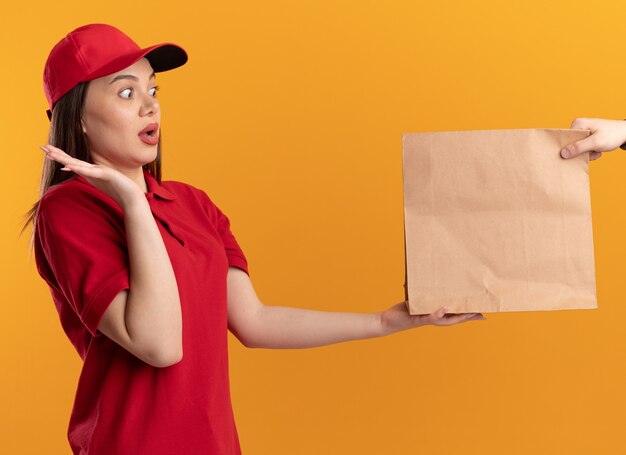 Anxious pretty delivery woman in uniform stands with raised hand and gives paper package to someone on orange
