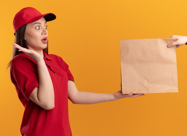 Anxious pretty delivery woman in uniform stands with raised hand and gives paper package to someone on orange