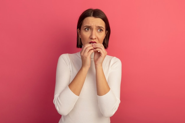 Anxious pretty woman bites nails looking at side isolated on pink wall