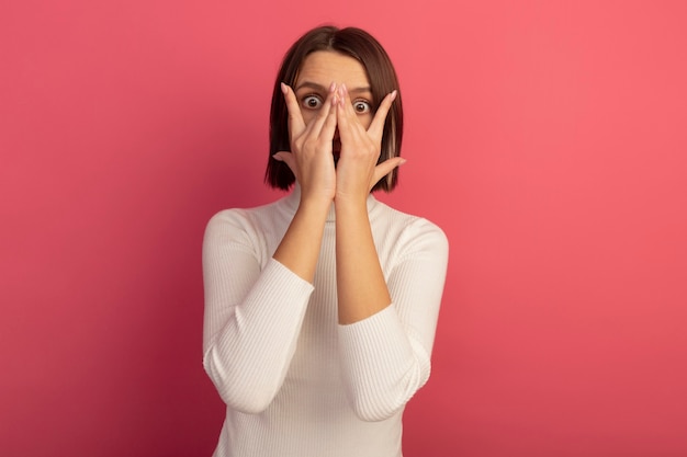 Free photo anxious pretty woman holds hands in front of face and looks at front through fingers isolated on pink wall