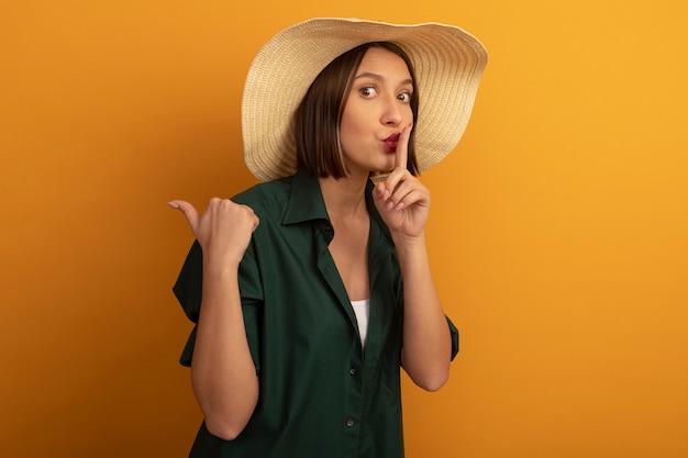 Обеспокоенная красивая женщина в пляжной шляпе делает жест молчания и указывает в сторону, изолированную на оранжевой стене