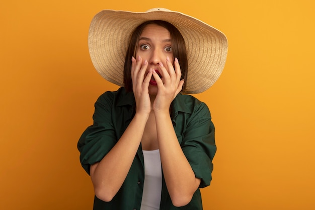 ビーチ帽子をかぶった気になるきれいな女性は、オレンジ色の壁で隔離の口の近くに手を保持します。