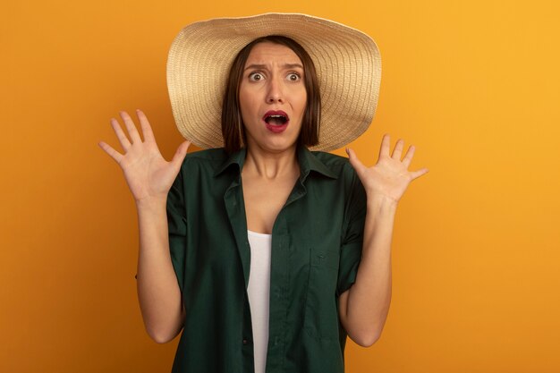 Обеспокоенная красивая женщина в пляжной шляпе стоит с поднятыми руками, изолированными на оранжевой стене