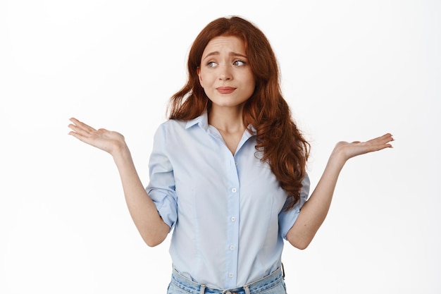 Anxious redhead woman shrugging shoulders indecisive, looking aside with panic confused look, dont know nothing, cant understand, have no clue, standing against white background