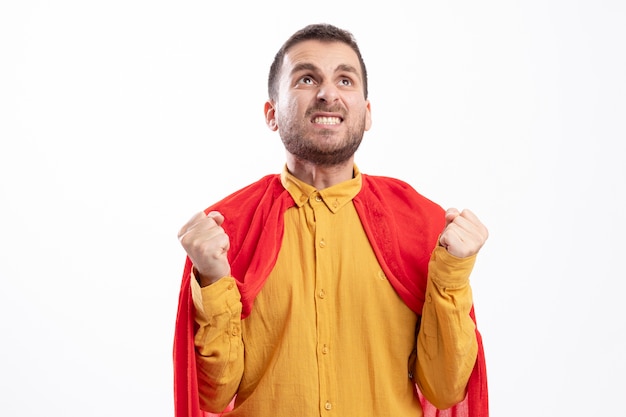 Anxious superhero man with red cloak keeps fists and looks up isolated on white wall