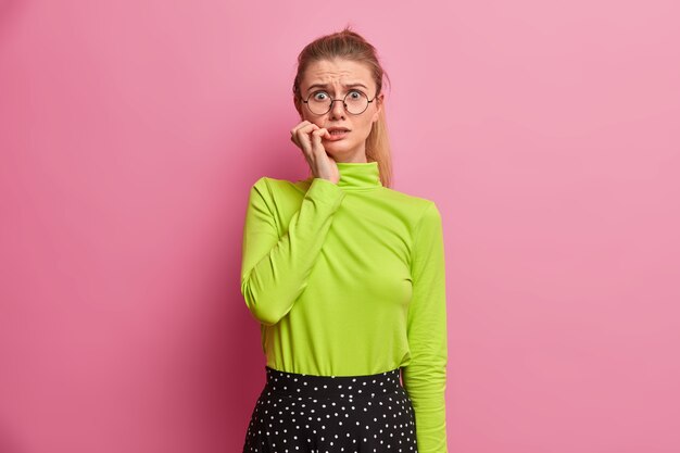 心配しているヨーロッパの女の子が指の爪を噛んで何かを恐れている、大きな問題を抱えている、緊張している、光学メガネをかけている、緑のタートルネック
